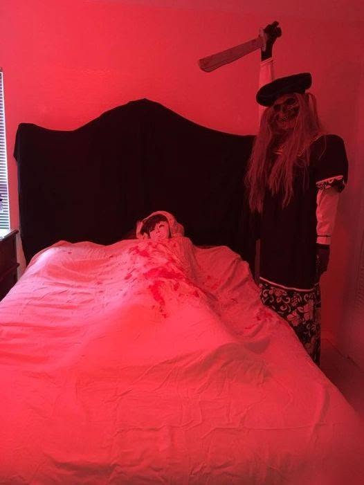 A red-lit bedroom scene with a figure in bed and a grim reaper-like prop standing beside it at The Witch House in 2018