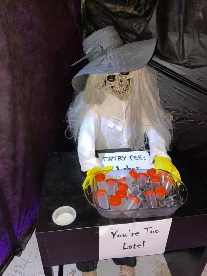 A skeletal attendant in a wide hat presenting sample cups and a sign at the 2020 Halloween party