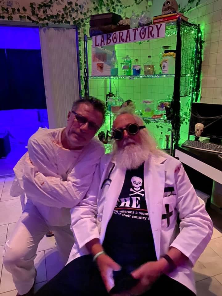 Two guests dressed as deranged medical staff posing in front of the laboratory display at the 2020 Halloween party