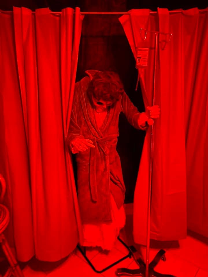 A red-lit figure emerging from behind hospital curtains with an IV stand at the 2020 Halloween party