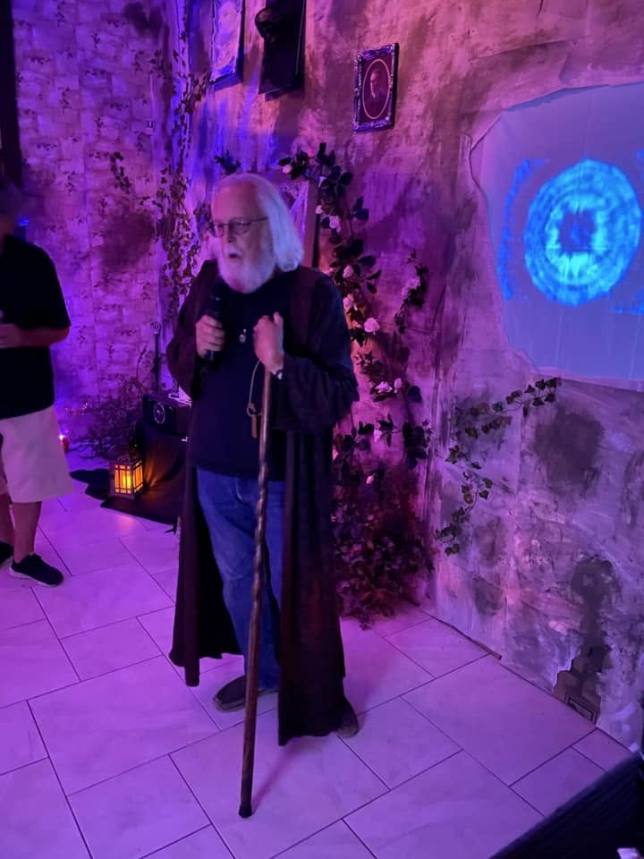 An elder robed figure holding a staff and speaking into a microphone at the 2024 party