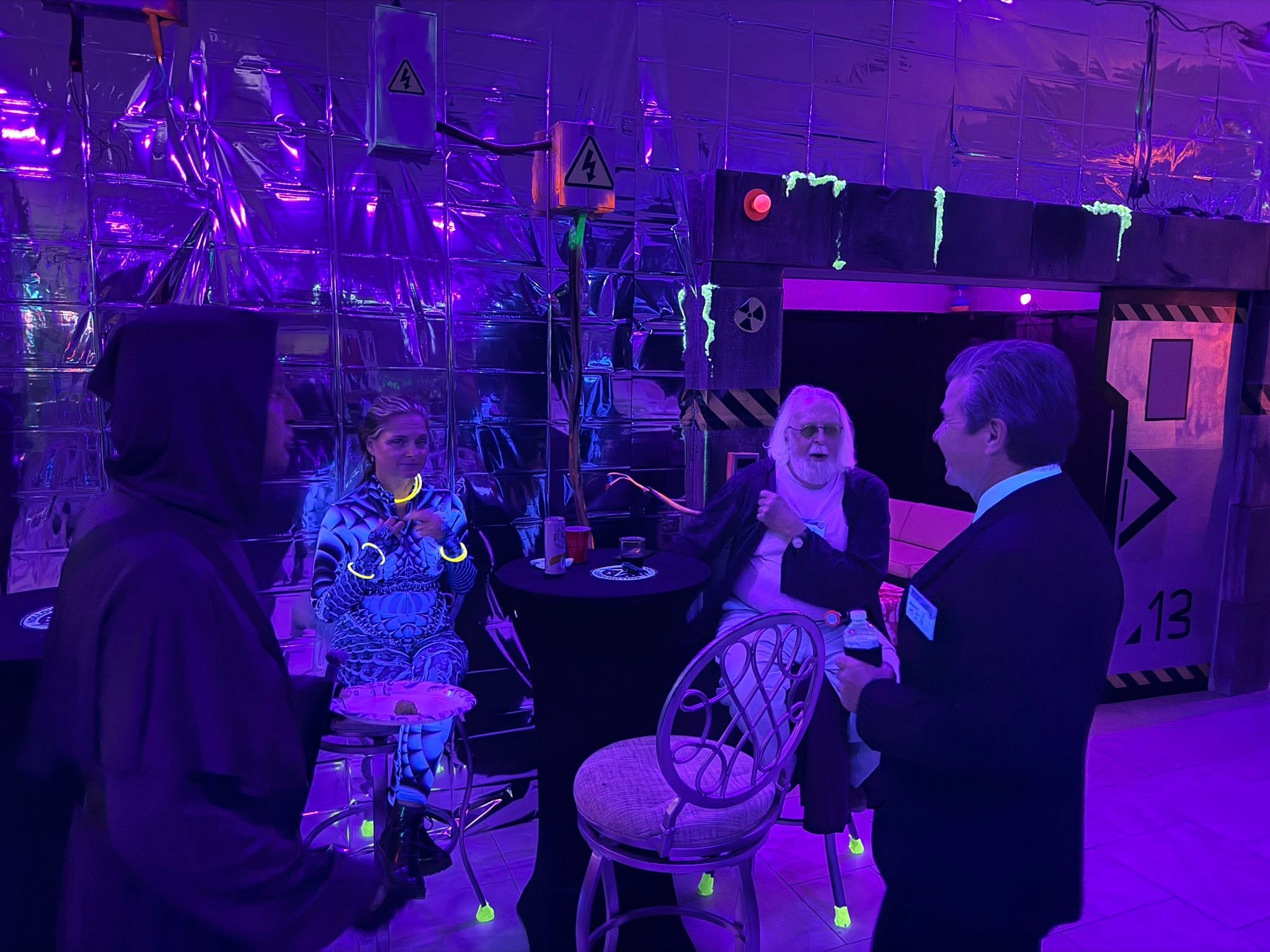 A small group of guests gathered in a neon purple space-station lounge beside a bay marked 13 at The Space Station in 2025