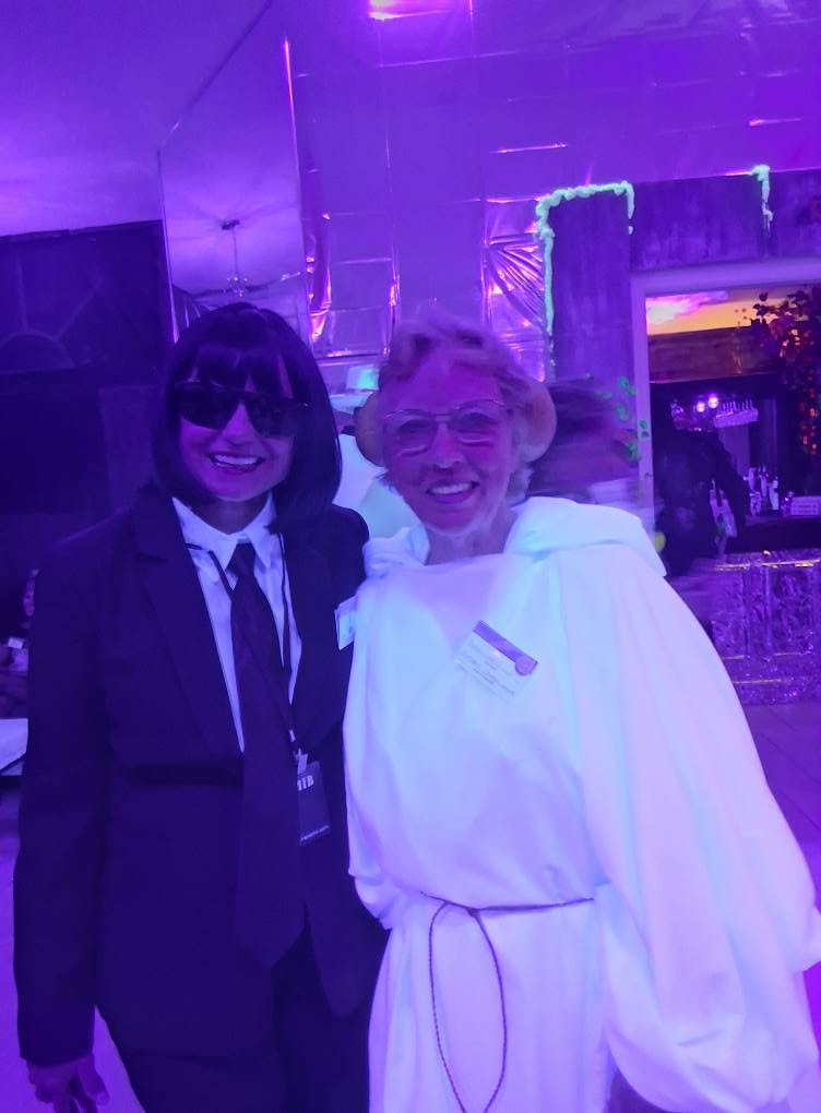 Two smiling guests posing together, one in a black agent suit and one in flowing white robes, at The Space Station in 2025