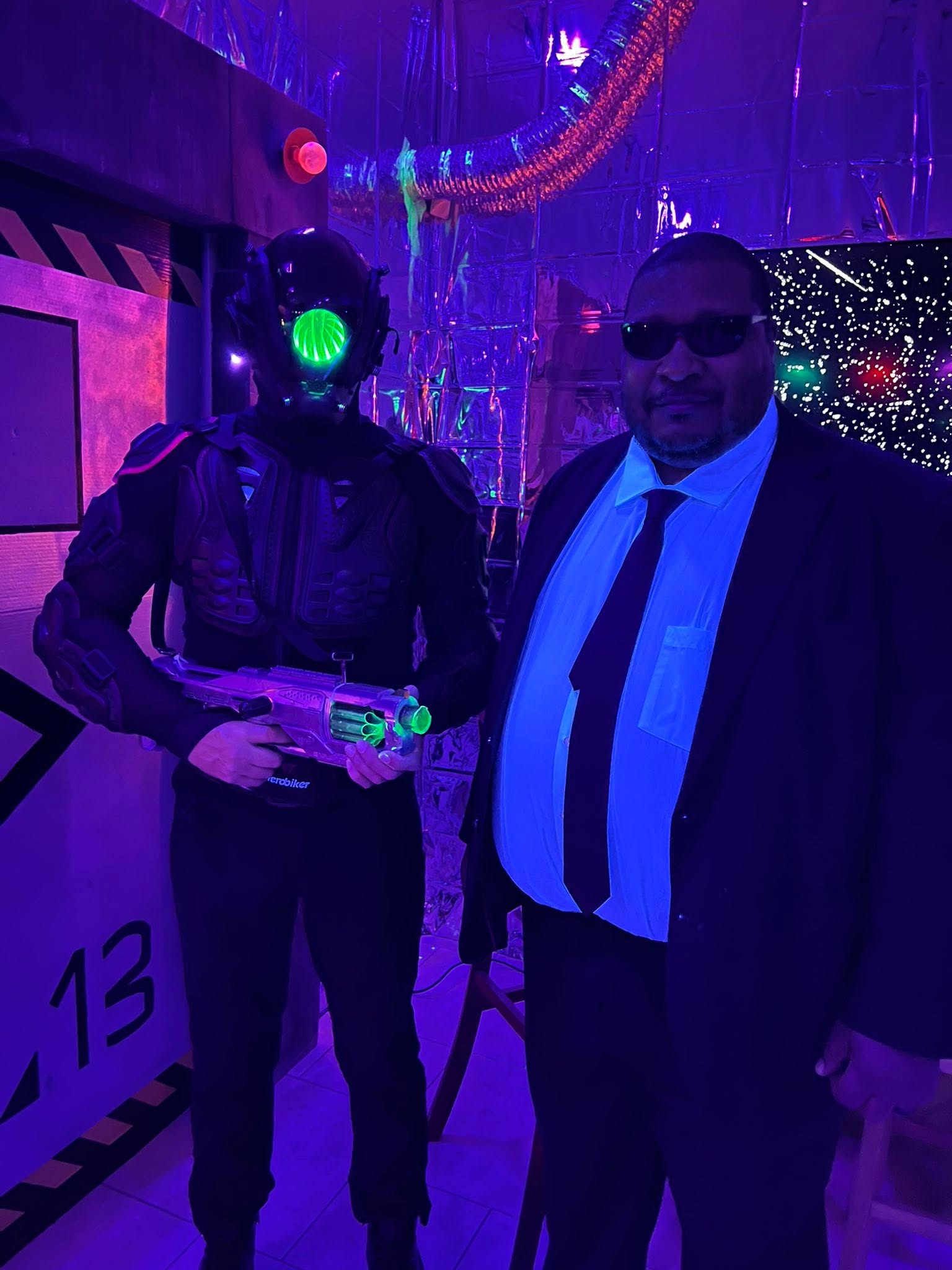 A futuristic armored guest with a glowing green visor posing beside a suited agent at The Space Station in 2025